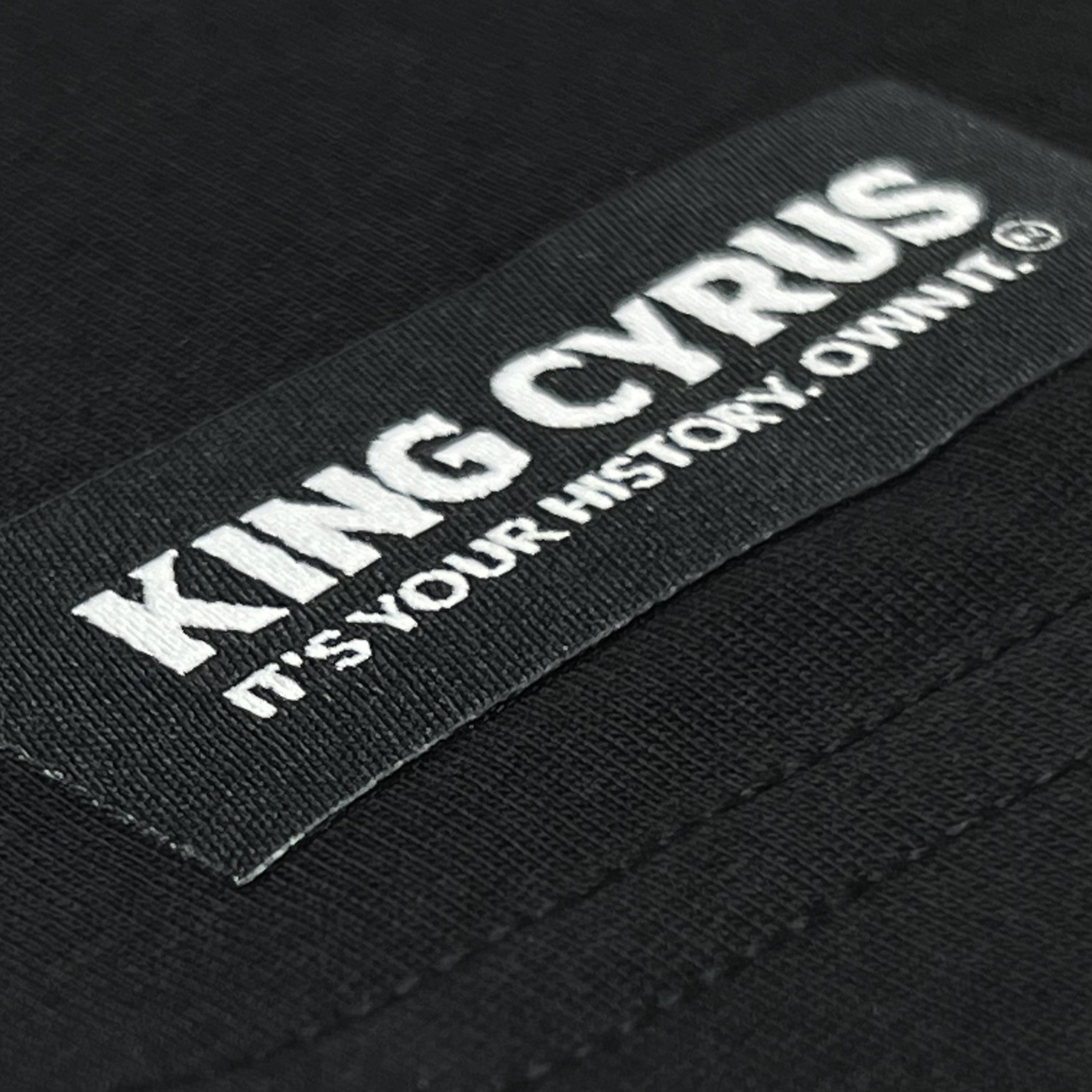 King Cyrus woven tag with tagline 'It’s Your History. Own It.' on front hem of black tee